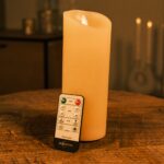 Die wiederaufladbare LED-Wachskerze DORSETTO (Ø 7,5 x 20 cm) steht auf einem Holztisch, daneben eine Fernbedienung; die Kerze leuchtet warm, im Hintergrund sind unscharf weitere Kerzen zu erkennen.