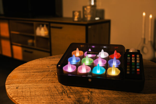 Auf einem runden Holztisch in einem warm beleuchteten Raum leuchten die LED-Teelichter DORSETTO wiederaufladbar (12 Stück) RGB mit Fernbedienung zwischen Kerzen, mit einem Holzschrank im Hintergrund.