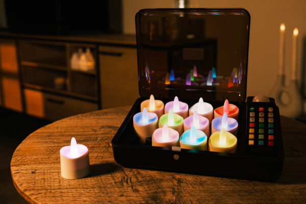 Auf einem Holztisch steht eine schwarze Box mit 12 wiederaufladbaren LED-Teelichtern DORSETTO (RGB), die jeweils in verschiedenen Farben leuchten. Eine bunte Fernbedienung liegt daneben, und eine Kerze ist außerhalb der Box platziert.