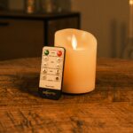 Eine wiederaufladbare LED-Wachskerze mit beweglicher Flamme D 7,5 x 10 cm - warmweiß steht auf einem Holztisch, angelehnt an eine Fernbedienung, mit warmem Umgebungslicht und unscharfem Mobiliar im Hintergrund.