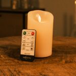 Eine wiederaufladbare LED-Wachskerze mit beweglicher Flamme D 7,5 x 12,5 cm (warmweiß) leuchtet auf einem Holztisch, davor steht eine Fernbedienung; im Hintergrund sorgt sanft-warmes Licht für stimmungsvolle Atmosphäre.