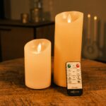 Zwei flammenlose LED-Kerzen unterschiedlicher Höhe stehen auf einem Holztisch und leuchten warm. Vor ihnen liegt eine Fernbedienung mit der Aufschrift Dorsetoo. Im Hintergrund sind verschwommene Kerzen und Möbel zu sehen.
