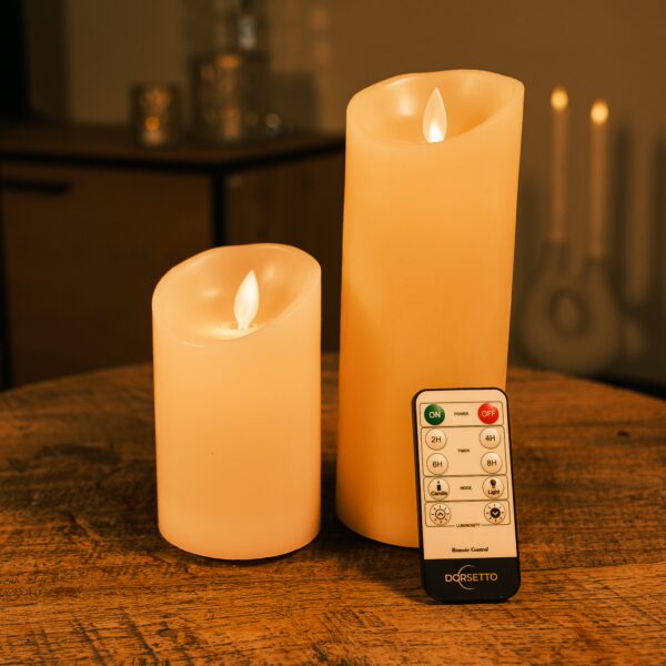 Twee Oplaadbare LED wax kaarsen set van 2 DORSETTO van verschillende hoogtes staan gloeiend op een houten tafel. Een DORSETTO-afstandsbediening ligt ervoor, met een zacht verlichte achtergrond voor extra sfeer.