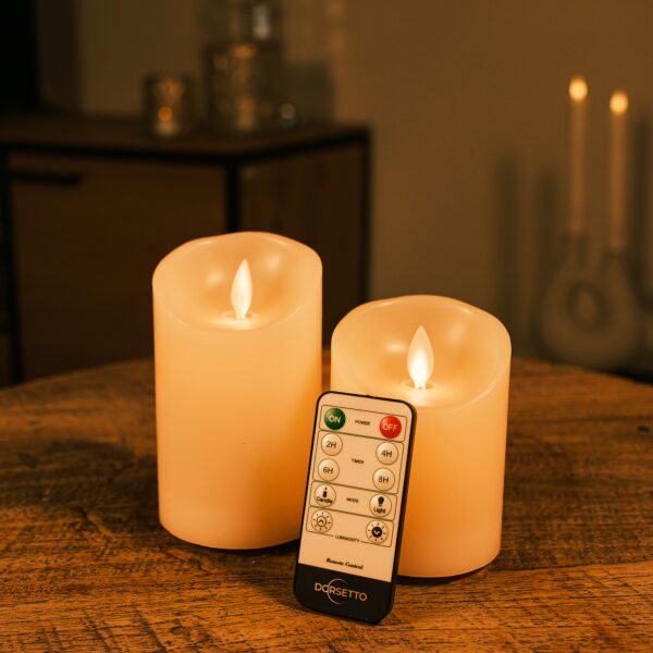 De LED kleine wax kaarsen set 2 kaarsen DORSETTO wordt weergegeven op een houten tafel met zijn afstandsbediening. Het warme LED-licht voegt een gezellige sfeer toe, terwijl op de achtergrond vage kaarsen en meubels te zien zijn.