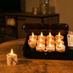 Op een ronde houten tafel staat een zwarte doos met daarin 12 3D oplaadbare LED-Waxine/theelichtjes (op batterijen, tot 400 uur) met timerfunctie en Afstandsbediening; één kaars staat buiten. Warme binnenverlichting maakt het tafereel compleet.