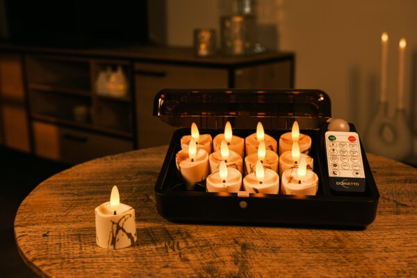 Op een ronde houten tafel staat een zwarte doos met daarin 12 3D oplaadbare LED-Waxine/theelichtjes (op batterijen, tot 400 uur) met timerfunctie en Afstandsbediening; één kaars staat buiten. Warme binnenverlichting maakt het tafereel compleet.