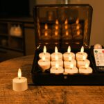 Ein Set aus zwölf flammenlosen LED-Teelichtern, die in einem schwarzen Ladekasten auf einem Holztisch stehen, wobei eine Kerze außerhalb des Kastens steht und eine Fernbedienung neben den Kerzen liegt.