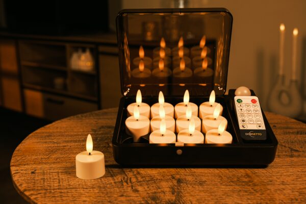 Ein Set aus zwölf flammenlosen LED-Teelichtern, die in einem schwarzen Ladekasten auf einem Holztisch stehen, wobei eine Kerze außerhalb des Kastens steht und eine Fernbedienung neben den Kerzen liegt.