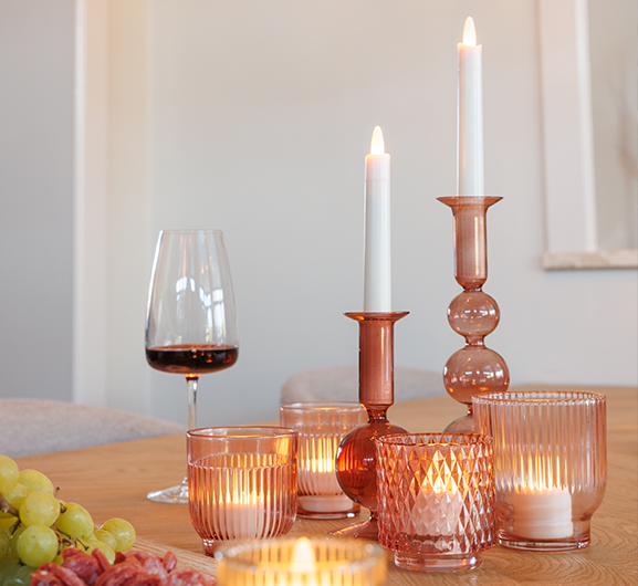 Ein Holztisch mit brennenden Kerzen in rosafarbenen Glashaltern, einem Glas Rotwein und einem Strauß grüner Weintrauben schafft eine warme, gemütliche Atmosphäre.