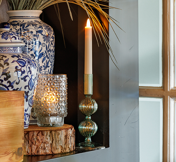 Eine brennende Kerze in einem Glashalter steht neben einem strukturierten Glasvotivhalter mit einer brennenden Kerze darin, beide auf einer Holzplatte, neben einer blau-weiß geblümten Keramikvase mit Ziergras.