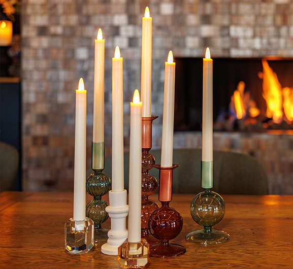 Sieben brennende Spitzkerzen in bunten und klaren Glashaltern sind auf einem Holztisch angeordnet, im Hintergrund brennt ein Kamin. Die Szene ist gemütlich und warm.