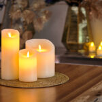 De LED wax kaarsen set 3 kaarsen warm wit oplaadbaar DORSETTO staat op een ronde geweven mat op een houten tafel, met wazige kaarsen en vazen op de achtergrond voor een warme, gezellige sfeer.
