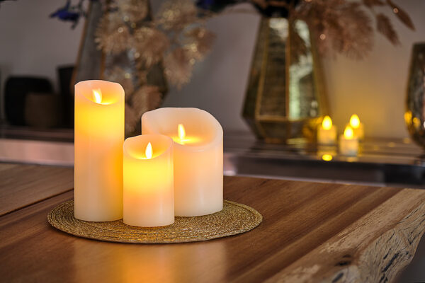 De LED wax kaarsen set 3 kaarsen warm wit oplaadbaar DORSETTO staat op een ronde geweven mat op een houten tafel, met wazige kaarsen en vazen op de achtergrond voor een warme, gezellige sfeer.