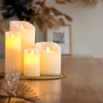 De Oplaadbare LED wax kaars met bewegende vlam D 7,5 X 10 CM - warm wit staat samen met andere kaarsen sfeervol op een gouden tray op een houten tafel, omringd door droog bladerwerk en een fotolijstje voor extra warmte en gezelligheid.