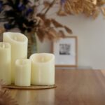 De LED wax kaarsen set 4 kaarsen warm wit oplaadbaar DORSETTO staat samen op een houten tafel, omringd door herfstbladeren met een vage ingelijste foto op de achtergrond.