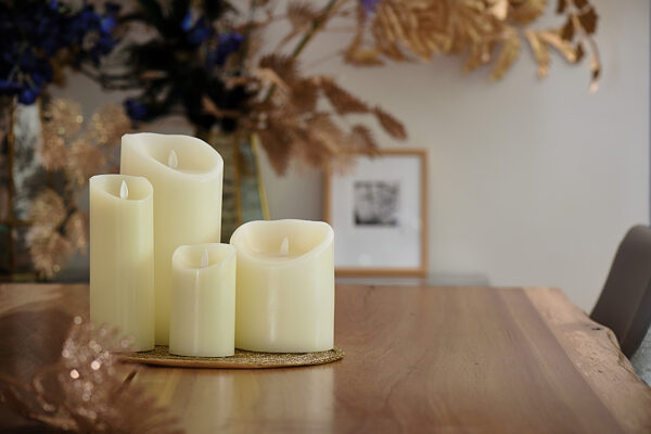 Vier Oplaadbare LED waxinelichtjes met bewegende vlam D 7,5 X 10 CM - warm wit in ivoor zijn gerangschikt op een houten tafel met gouden accenten. De onscherpe achtergrond is voorzien van gedroogd gebladerte en ingelijste kunst voor een elegante, gezellige sfeer.