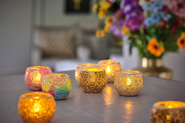 Zeven Waxinelichthouder 9 CM Dorsetto mozaïek multicolor glazen waxinelichthouders gloeien in levendige kleuren op een tafel, terwijl vage kleurrijke bloemen en een bank op de achtergrond bijdragen aan de gezellige, decoratieve sfeer.