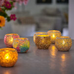 Zes decoratieve mozaïek kandelaars met LED- waxine/theelichtjes waterdicht oplaadbaar (warm wit, 175 uur brandtijd, 12 stuks, met afstandsbediening) staan op een tafel; drie zijn goudkleurig en drie zijn veelkleurig. Bloemen en neutrale meubels vervagen op de achtergrond.