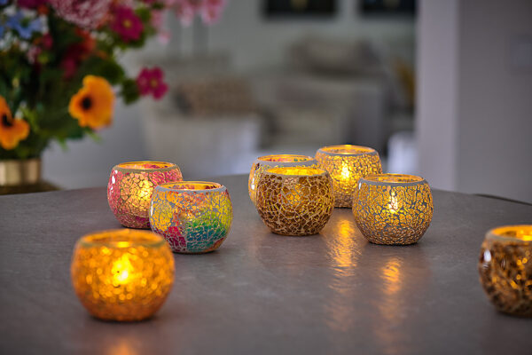 Twaalf waterdichte, oplaadbare LED-theelichtjes (brandduur 175 uur, warm wit) met afstandsbediening gloeien in mozaïekglazen houders met gouden en kleurrijke patronen. Op de onscherpe achtergrond staan bloemen en een gezellige woonkamer.