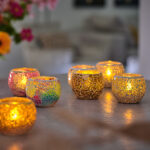 De Waxinelichthouder 8,5 CM Dorsetto mozaïek rood, met een brandende kaars erin, staat uitgestald tussen andere kleurrijke mozaïek glazen houders op een tafel. Op de zacht vervaagde achtergrond staan bloemen en een warme, gezellige omgeving binnenshuis.