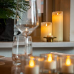 Close-up van een gedekte tafel met lege wijn- en waterglazen, met de LED wax kaarsen set 2 maten warm met oplaadbaar DORSETTO en kaarsen op de tafel en vensterbank voor een gezellige sfeer, met groen decor op de achtergrond.