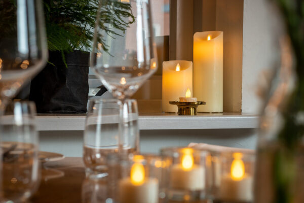 Close-up van een gedekte tafel met lege wijn- en waterglazen, met de LED wax kaarsen set 2 maten warm met oplaadbaar DORSETTO en kaarsen op de tafel en vensterbank voor een gezellige sfeer, met groen decor op de achtergrond.