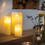 De LED wax kaarsen set 3 kaarsen warm wit oplaadbaar DORSETTO, met kaarsen van verschillende hoogtes, staat op een houten oppervlak tegen een muur met witte structuur. Zachte verlichting en vage glazen potten met bloemen dragen bij aan de gezellige sfeer.