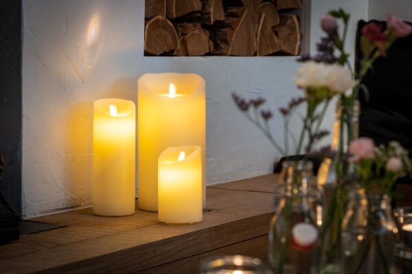 De LED wax kaarsen set 3 kaarsen warm wit oplaadbaar DORSETTO, met kaarsen van verschillende hoogtes, staat op een houten oppervlak tegen een muur met witte structuur. Zachte verlichting en vage glazen potten met bloemen dragen bij aan de gezellige sfeer.