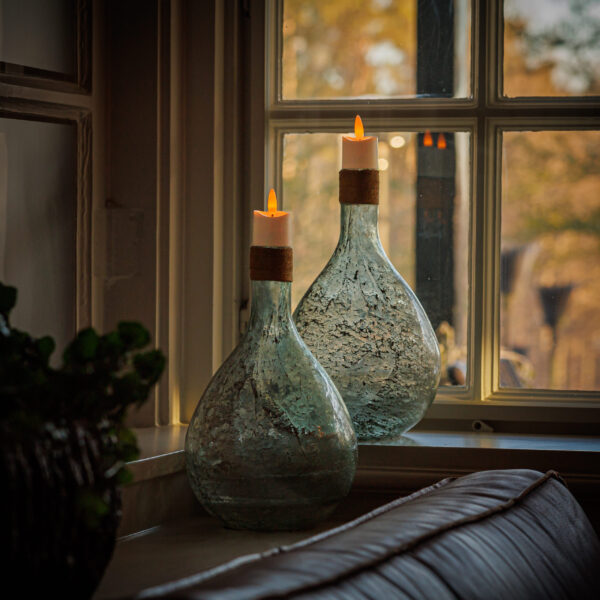 Op een vensterbank staan twee glazen vazen met daarop 3D oplaadbare LED-thee/waxinelichtjes 175 uur (set van 6, met afstandsbediening en timerfunctie) die zacht oplichten als het zonlicht naar binnen stroomt; een plant en een deel van een bank zijn vlakbij zichtbaar.