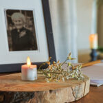 Een ingelijst zwart-wit portret, kleine witte bloemen en een 3D oplaadbaar LED-thee/waxinelichtje (175 uur, timerfunctie, met afstandsbediening, set van 6) rusten op een houten plaat, wat een gedenkteken suggereert. Op de achtergrond zijn een vage kaars en een boek zichtbaar.