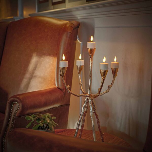 Een gouden kandelaarlamp met vijf brandende gloeilampen staat op een houten tafel naast een bruine leren fauteuil in een gezellige kamer. LED waxinelichtjes 3D (12 stuks, oplaadbaar met afstandsbediening) en een kleine potplant geven de ruimte extra charme.