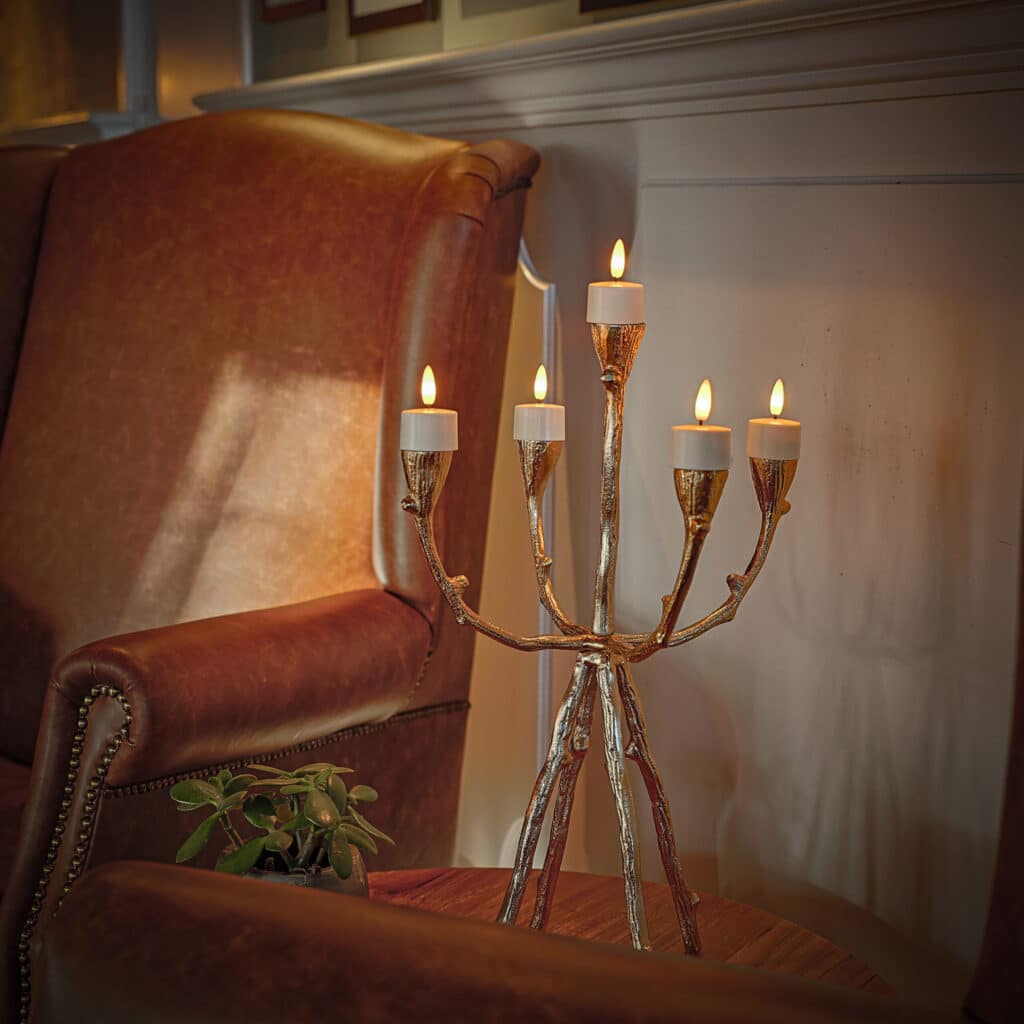 Een gouden kandelaar met zes LED waxinelichtjes 3D (brandtijd 175 uur, oplaadbaar, afstandsbediening, timerfunctie) staat op een houten tafel tussen twee bruinleren fauteuils, met een kleine plant vlakbij en witte lambrisering erachter.