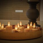 Een rond dienblad toont zes 3D oplaadbare LED-was/theelichtjes (175 uur, met afstandsbediening, timerfunctie), hun warme gele licht creëert een gezellige sfeer. Twee donkere kaarsenhouders staan op de achtergrond op een glad oppervlak.