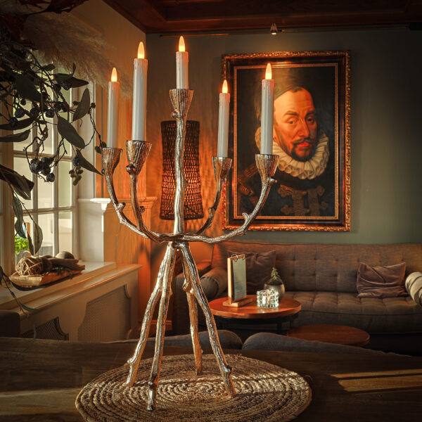 Een zilveren kandelaar met zes LED dinerkaars oplaadbaar 97mm 130 uur warm wit (set van 6, met afstandsbediening) staat op een geweven mat in een gezellige kamer en werpt warm licht bij een bank en een groot ingelijst portret.