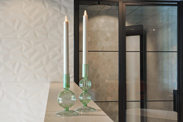 Twee hoge witte LED dinerkaarsen 270mm oplaadbaar uit een 6-delige set met afstandsbediening, die 130 uur lang warm wit opgloeien, staan in elegante groene houders op een licht aanrecht, tegen geometrische witte tegels en een glazen deur.