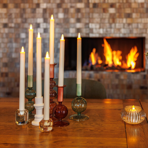Verschillende brandende kaarsen in kleurrijke glazen houders en een kleine geribbelde kaars staan op een houten tafel. Een Oplaadbare LED dinerkaars 270mm 130 uur warm wit gloeit vlakbij, terwijl de open haard bijdraagt aan de gezellige sfeer.