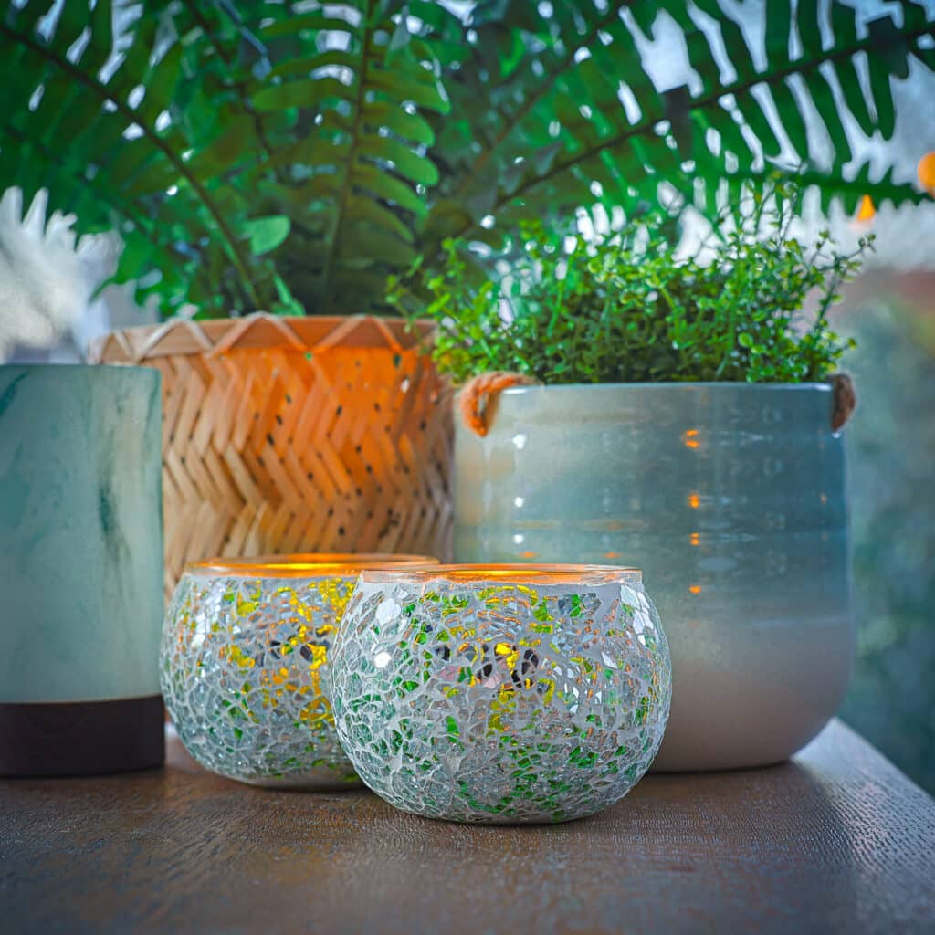 Twee mozaïek glazen kandelaars met LED waxine lichtjes oplaadbaar met afstandsbediening waterdicht brandtijd 175 uur gloeien op een tafel naast een groene potplant in een geweven mand en een andere in een blauwe pot van keramiek, met een zacht wazige achtergrond die warmte toevoegt.