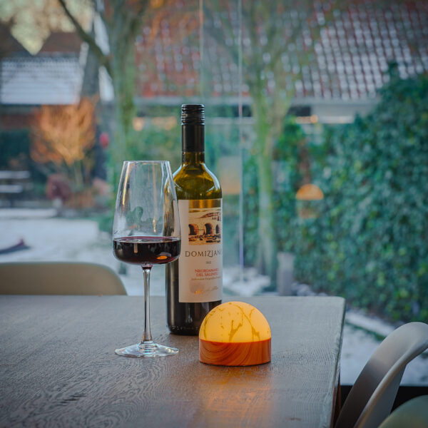 Een fles rode wijn, een gevuld glas en het Tafellamp DORSETTO oplaadbaar 105 uur waterdicht ovaal marmer groen houtlook grijs goud met afstandsbediening gloeien op een houten tafel bij het raam, met uitzicht op een besneeuwde tuin en huizen in de verte.