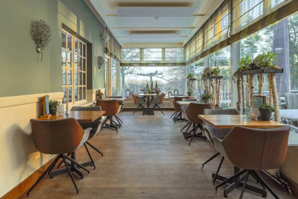 Een lichte, gezellige eethoek met houten vloeren, moderne stoelen en kleine tafels. Grote ramen laten natuurlijk licht binnen en bieden uitzicht op bomen. Planken en tafels zijn versierd met groene planten en 3D oplaadbare LED-theelichtjes (set van 12) met timerfunctie.