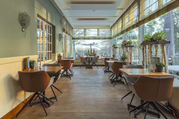 Een gezellig, zonovergoten café met houten vloeren, moderne bruine stoelen en kleine tafeltjes voor twee wordt zacht verlicht door LED waxine lichtjes oplaadbaar met afstandsbediening waterdicht brandtijd 175 uur, met potplanten en uitzicht op bomen buiten.