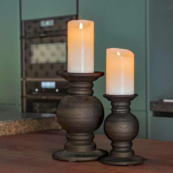 Twee Oplaadbare LED wax kaarsen set van 2 DORSETTO vlamloze kaarsen op donkere houders staan op een houten tafel, met een vage moderne keuken en ingebouwde oven op de achtergrond.