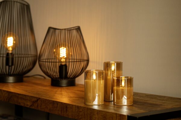 Drie DORSETTO oplaadbare led kaarsen (set van 3, brandtijd 105 uur, incl. afstandsbediening) in glazen houders staan op hout naast metalen lampen-perfect voor hotels en direct warme sfeer na opladen.