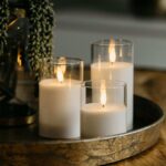 Drie DORSETTO led kaarsen (oplaadbaar, 105 uur, 3D vlam) staan verlicht in glazen houders op een rond goudkleurig dienblad. De afstandsbediening zit erbij en een groene plant is deels zichtbaar op de achtergrond.
