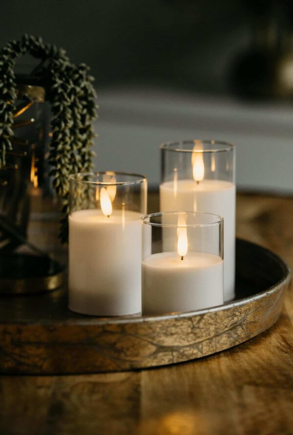 Drie DORSETTO led kaarsen (oplaadbaar, 105 uur, 3D vlam) staan verlicht in glazen houders op een rond goudkleurig dienblad. De afstandsbediening zit erbij en een groene plant is deels zichtbaar op de achtergrond.