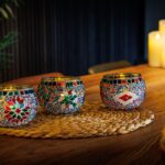 Drie levendige Waxinelichthouder 9 CM Dorsetto regenboog mozaïek glazen houders zijn aangestoken en gerangschikt op een geweven mat op een houten tafel, met pilaarkaarsen die gloeien op de achtergrond om een gezellige, warme sfeer te creëren.