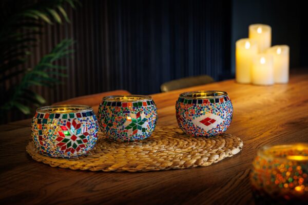Drie levendige Waxinelichthouder 9 CM Dorsetto regenboog mozaïek glazen houders zijn aangestoken en gerangschikt op een geweven mat op een houten tafel, met pilaarkaarsen die gloeien op de achtergrond om een gezellige, warme sfeer te creëren.