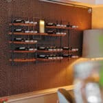 Een modern wijnrek aan de muur toont verschillende wijnflessen en een grote kaars boven een eettafel, aangevuld met de DORSETTO LED kleine was kaarsen set 2 kaarsen voor extra sfeer tegen een bruine muur met patroon.