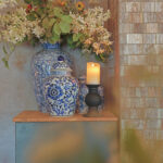 Op een houten tafel staan sierlijke blauw-witte bloemenvazen - één met bloemen en groen - naast een Oplaadbare LED wax kaars met bewegende vlam D 7,5 X 10 CM - warm wit in een zwarte houder, tegen een betegelde muur.