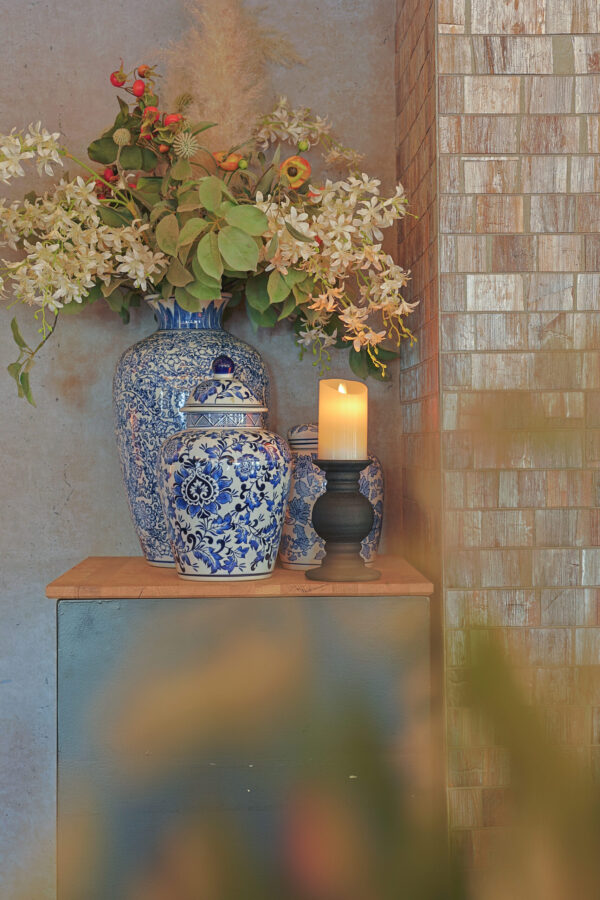 Op een houten tafel staan sierlijke blauw-witte bloemenvazen - één met bloemen en groen - naast een Oplaadbare LED wax kaars met bewegende vlam D 7,5 X 10 CM - warm wit in een zwarte houder, tegen een betegelde muur.
