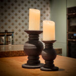 Twee Oplaadbare LED wax kaarsen met bewegende vlam D 7,5 X 10 CM - warm wit staan op donkere, rustieke kandelaars op een houten oppervlak in een warm verlichte keuken met patroonbehang.